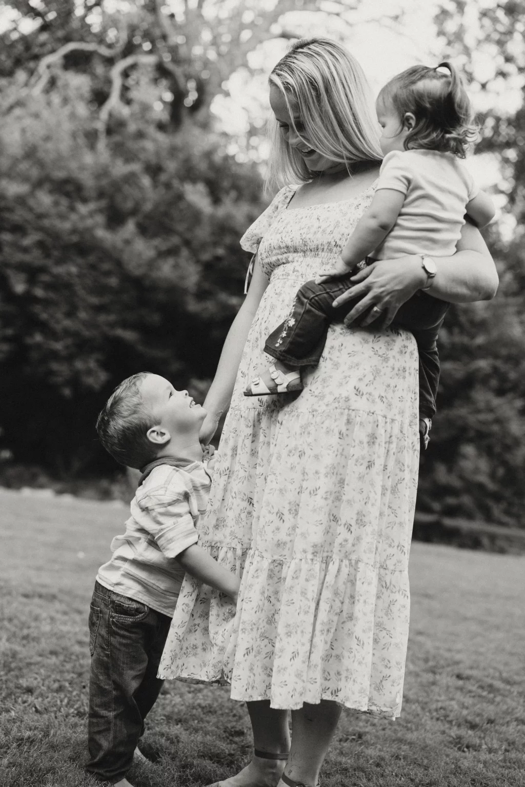 Family session at the Rock Quarry Garden.: Black-and-white portrait of a mother in a floral dress holding a toddler while a young boy hugs her leg outdoors.