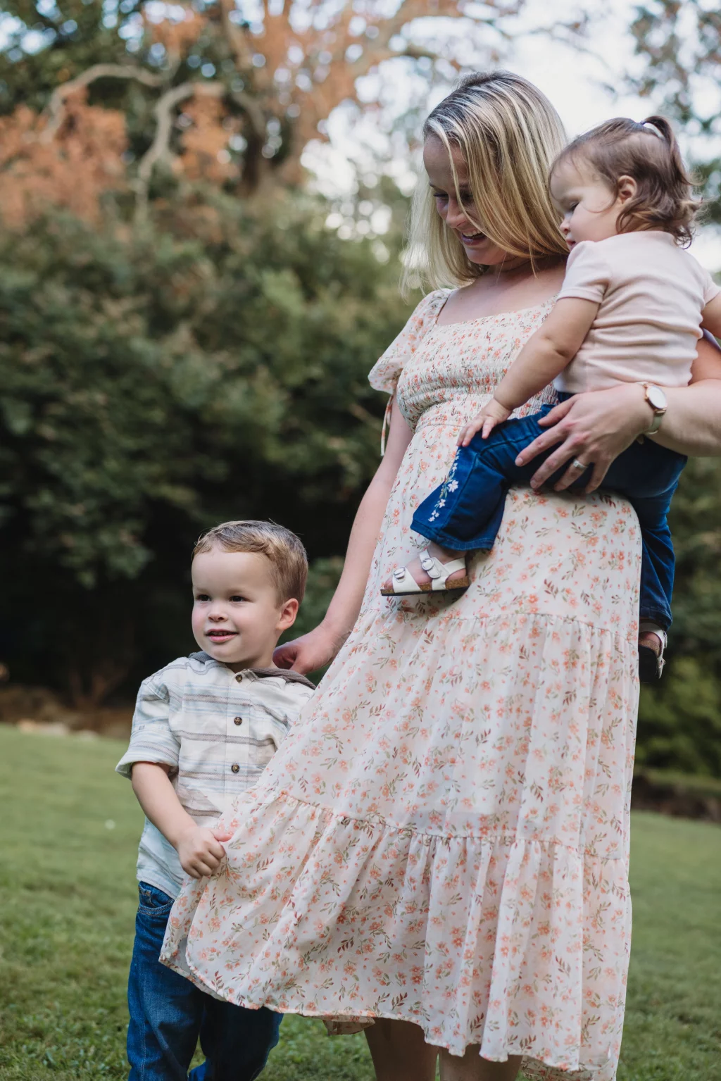 Family session at the Rock Quarry Garden.: A smiling mother in a floral dress holds a baby while a young boy stands beside her in a grassy, wooded park.