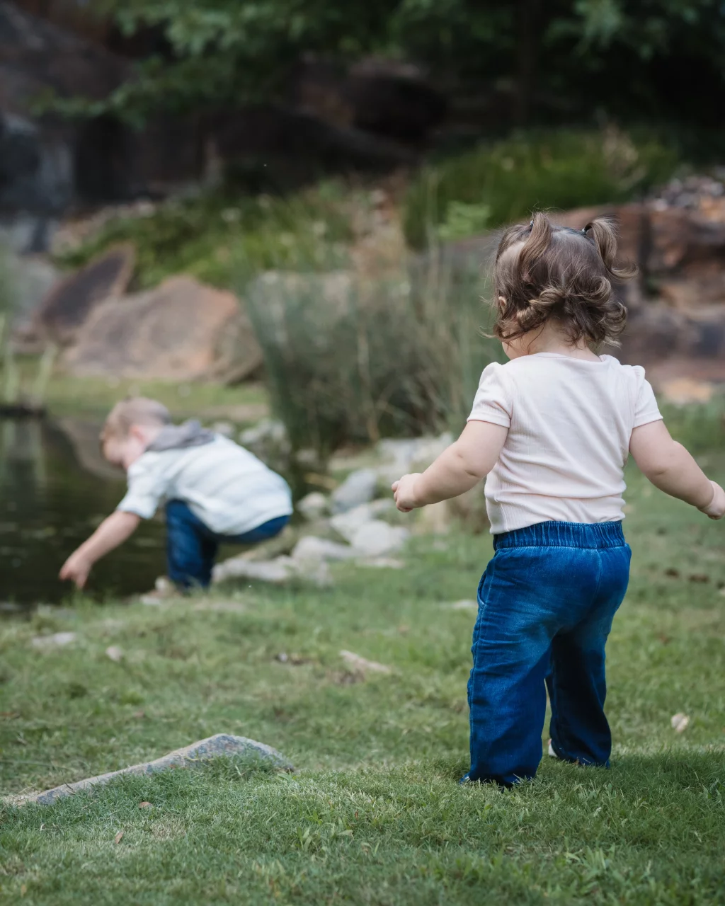 Family session at the Rock Quarry Garden.: Two young children explore a grassy, rocky area beside a pond; a toddler with pigtails stands in the foreground while another child crouches near the water.