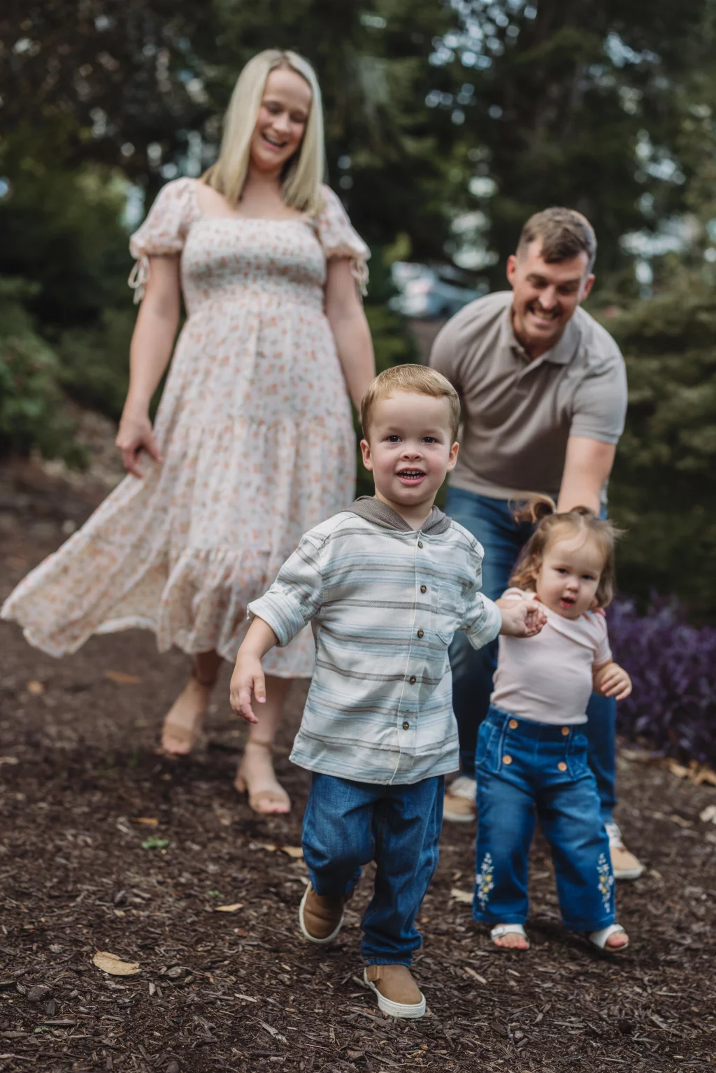 Family session at the Rock Quarry Garden.: A smiling family walks through a wooded garden, with a young boy in front holding hands with a toddler and their parents behind them.