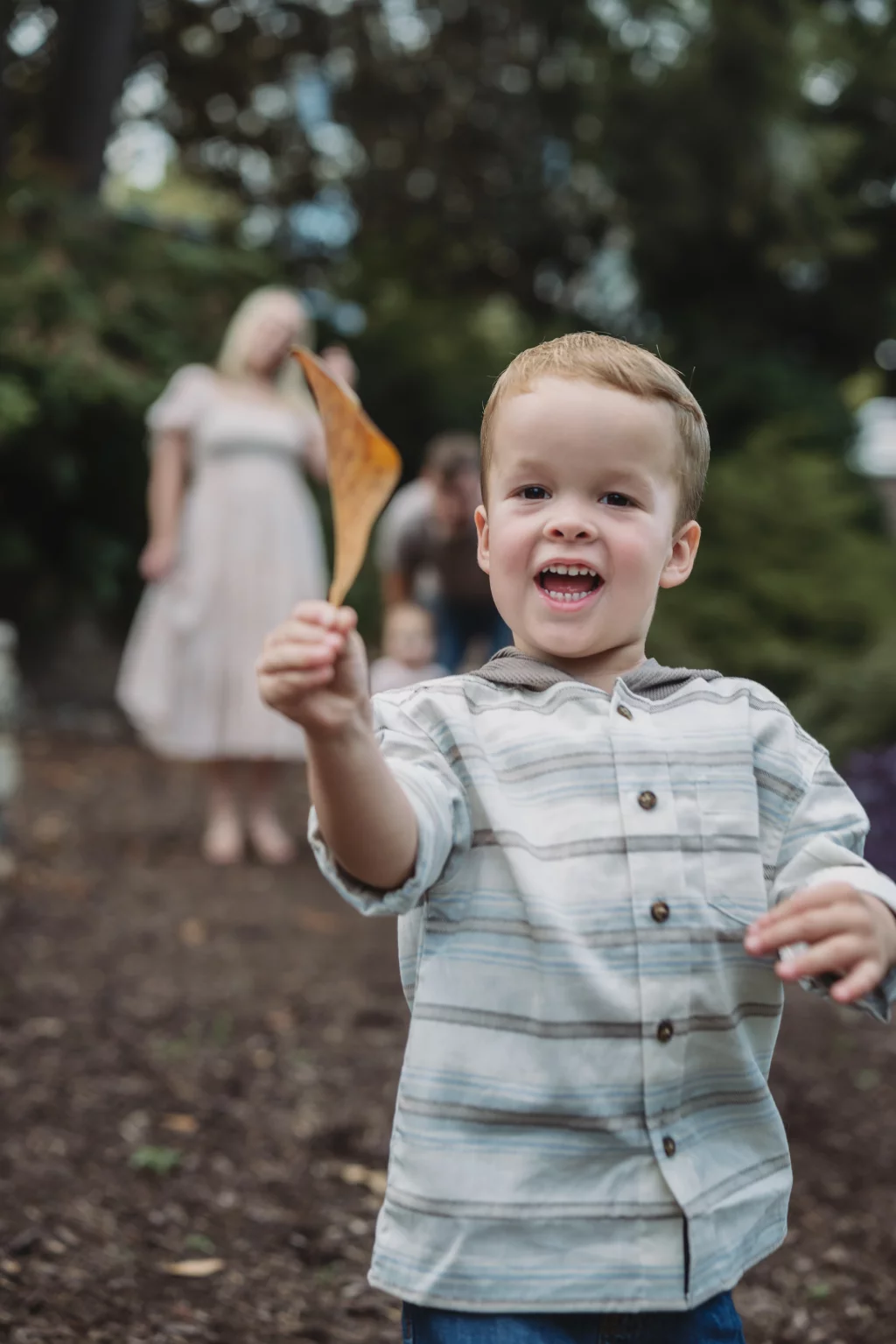 Family session at the Rock Quarry Garden.: Smiling young boy in a striped shirt holds up a brown leaf in a wooded park, with blurred adults in the background.