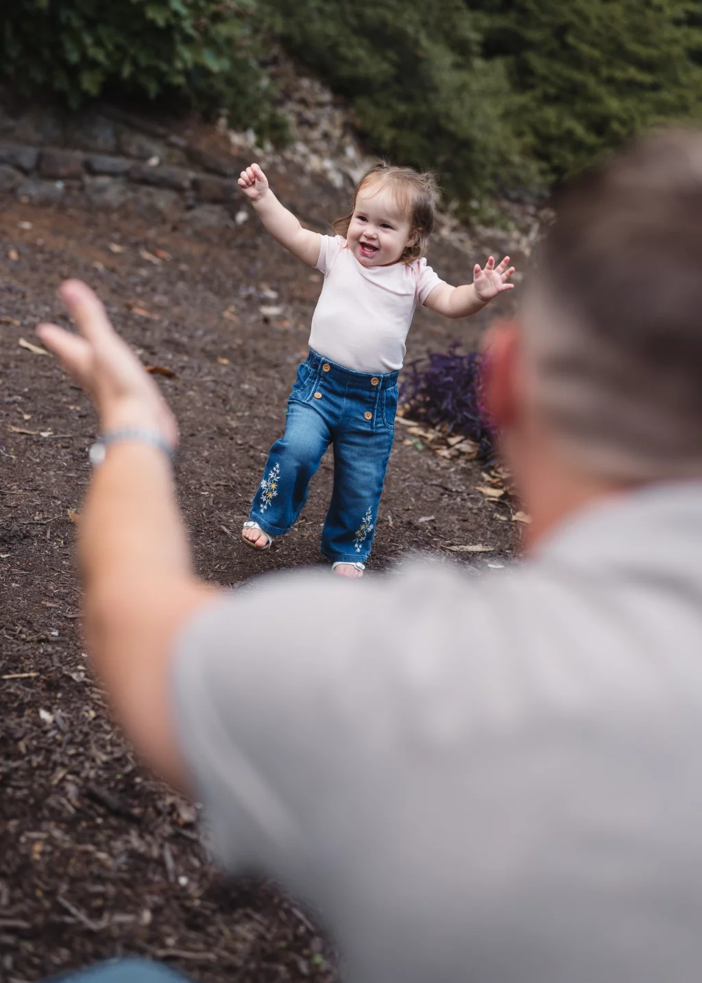 Family session at the Rock Quarry Garden.: Smiling toddler in a pink shirt and embroidered jeans raises her arms toward an adult in the foreground on a wooded path.