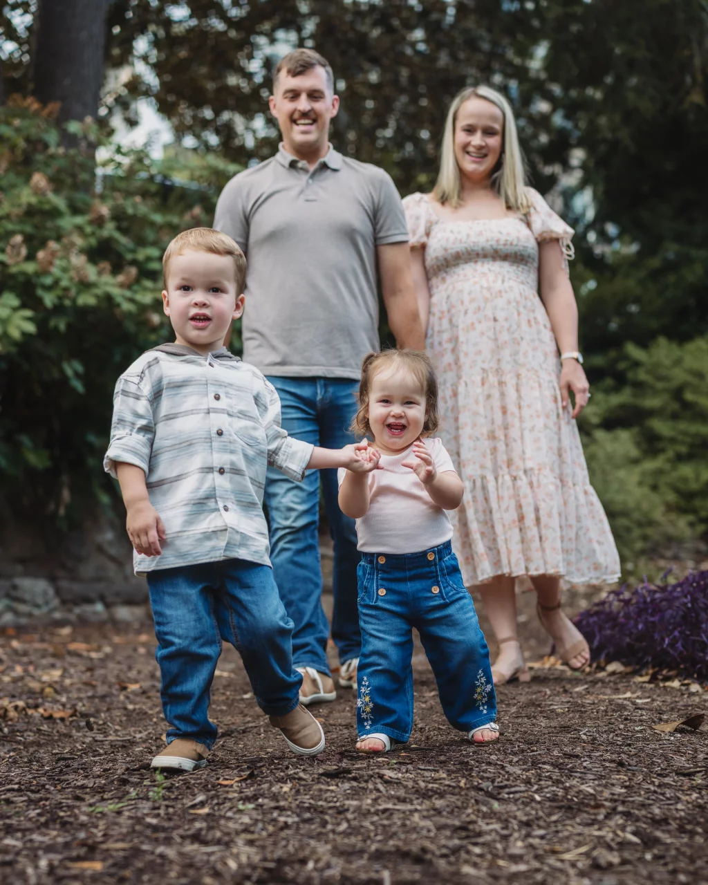 Family session at the Rock Quarry Garden.: A smiling family of four poses outdoors in a wooded garden, with two young children in front and their parents standing behind.