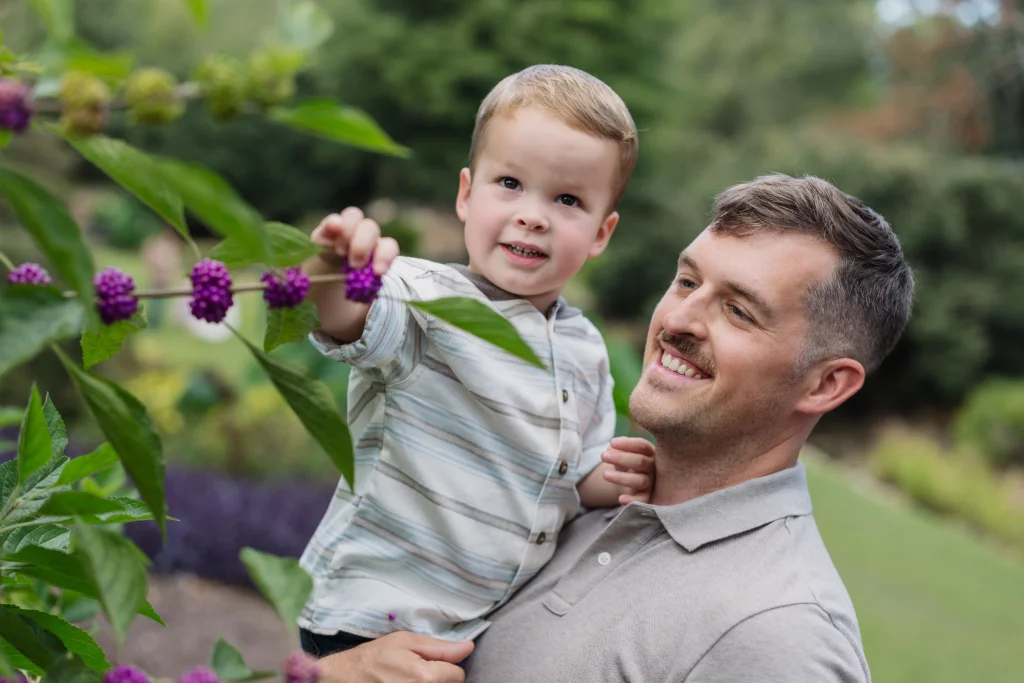 Family session at the Rock Quarry Garden.: A smiling man holds a young child outdoors as the child reaches toward purple flowers on a leafy branch.