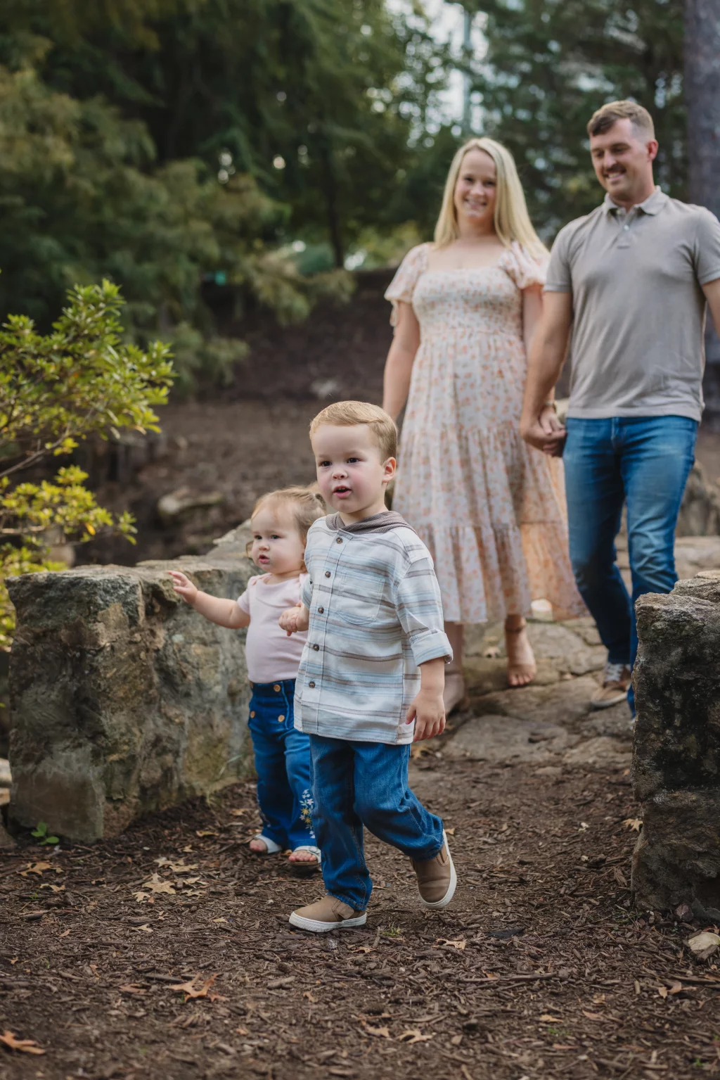 Family session at the Rock Quarry Garden.: A family walks along a wooded path between stone walls, with two young children in front and their parents holding hands behind them.