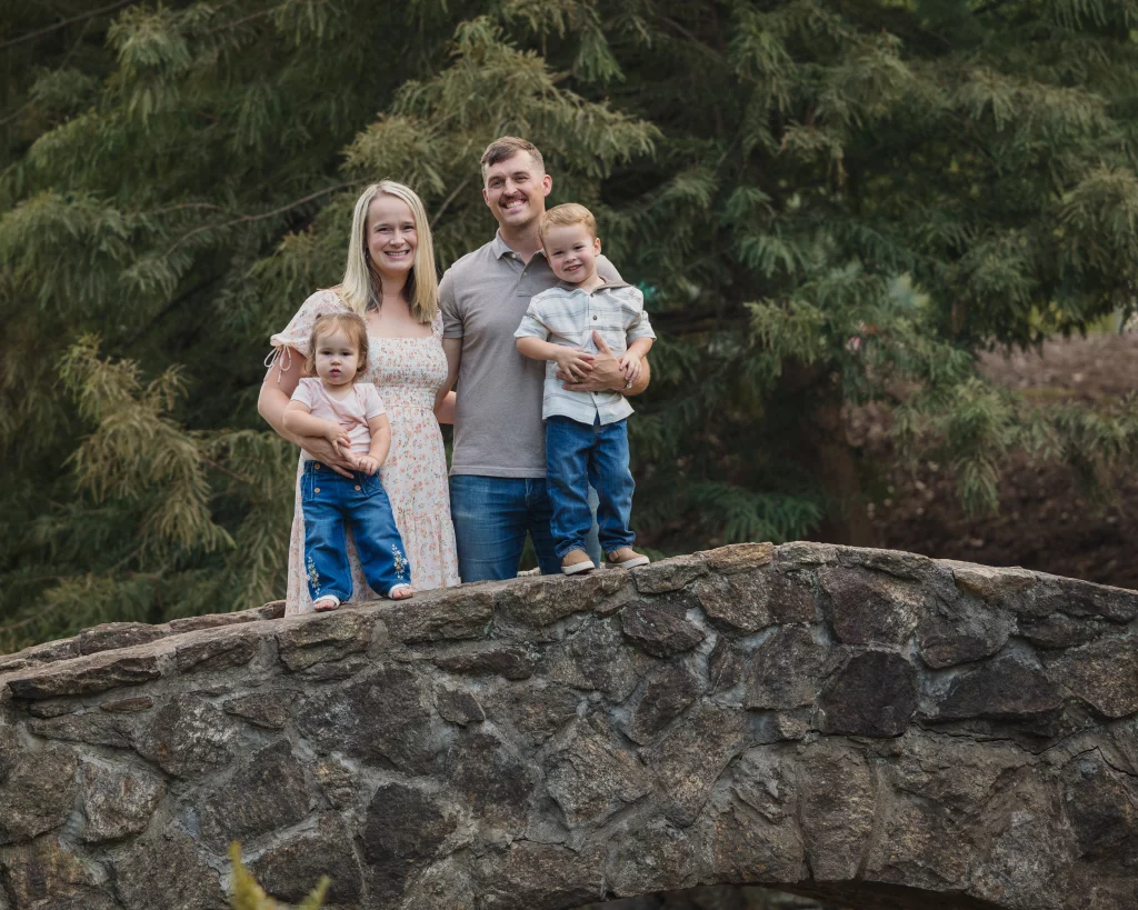 Family session at the Rock Quarry Garden.: A smiling family of four poses on a stone bridge in front of dense evergreen trees; the mother holds a toddler, while the father holds a young boy.