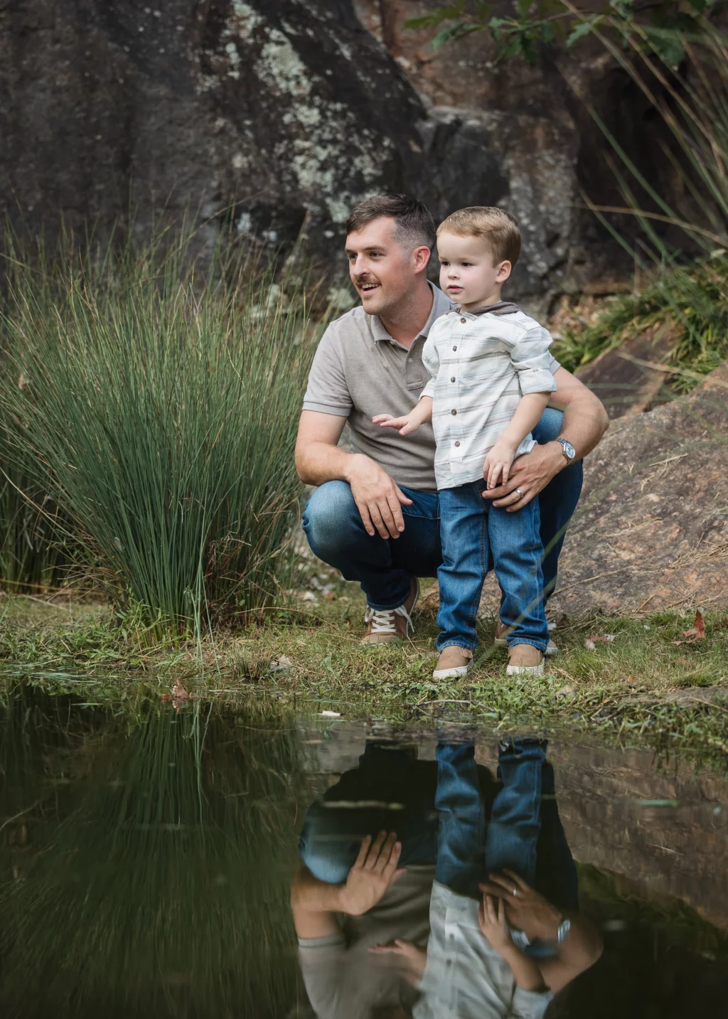 Family session at the Rock Quarry Garden.: A man kneels beside a pond holding a young boy, with rocks and tall grasses behind them and their reflections visible in the water.