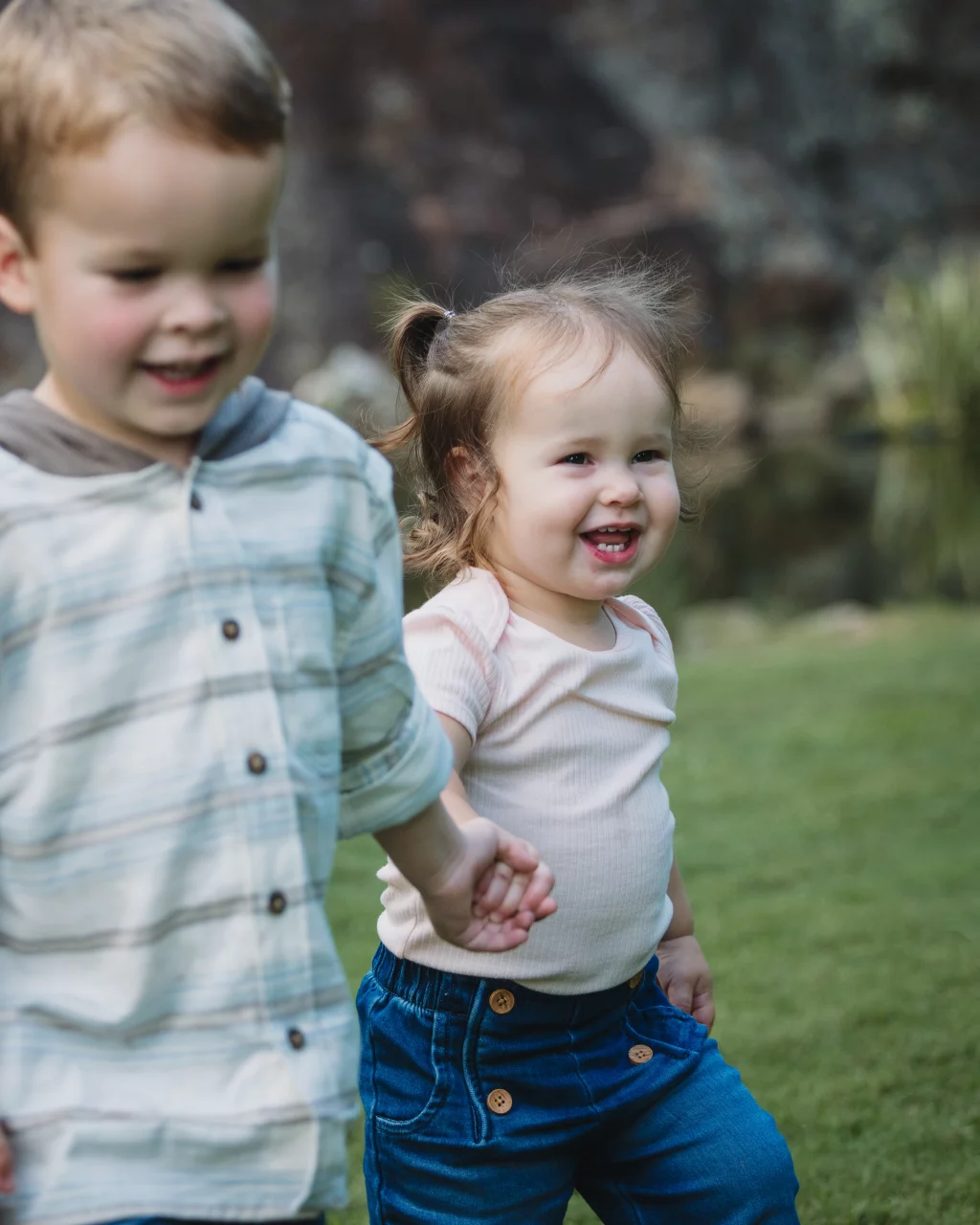 Family session at the Rock Quarry Garden.: Two young children smile while holding hands outdoors on a grassy lawn.