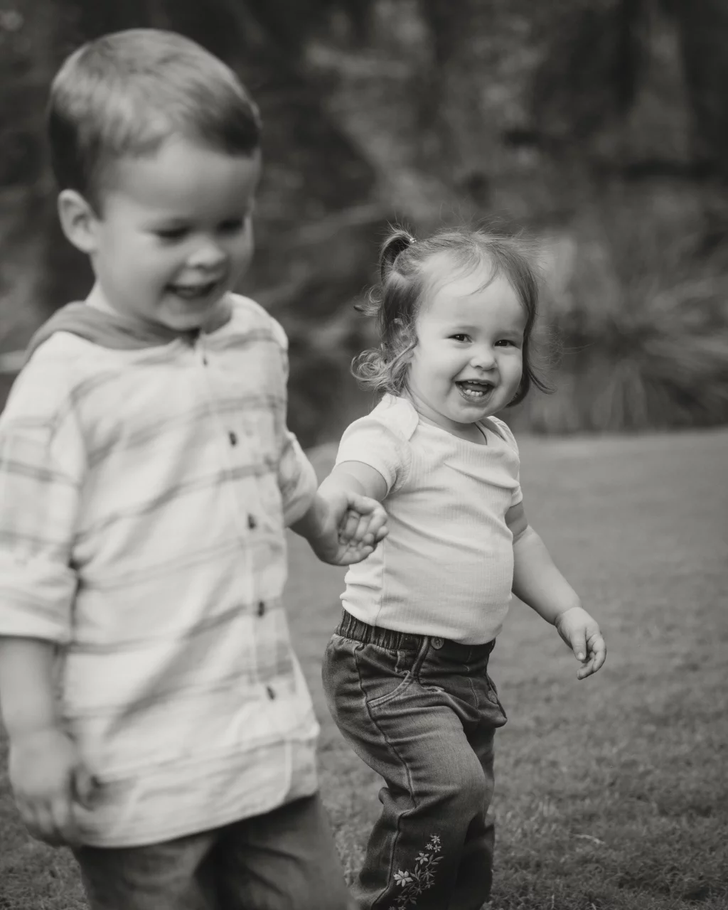 Family session at the Rock Quarry Garden.: Black-and-white photo of two smiling toddlers walking hand in hand outdoors on a grassy lawn.