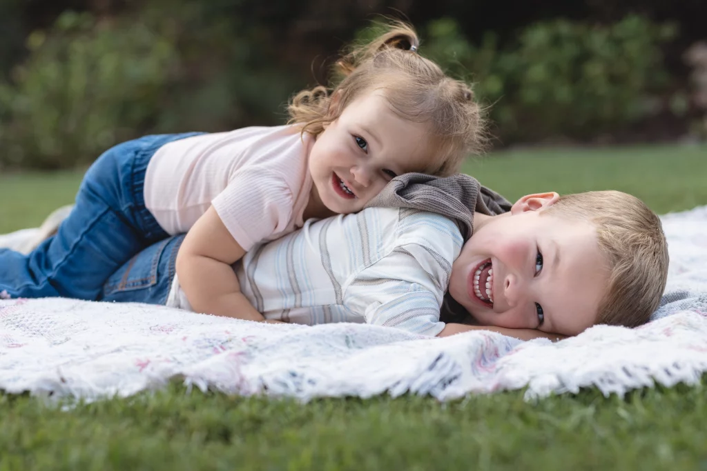 Family session at the Rock Quarry Garden.: Two smiling children playfully lie on top of each other on a blanket in a grassy park.