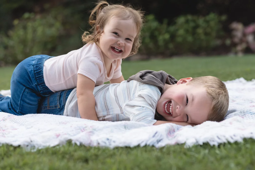 Family session at the Rock Quarry Garden.: Two smiling children play on a blanket in a grassy yard, with the younger child sitting on the older child&rsquo;s back.