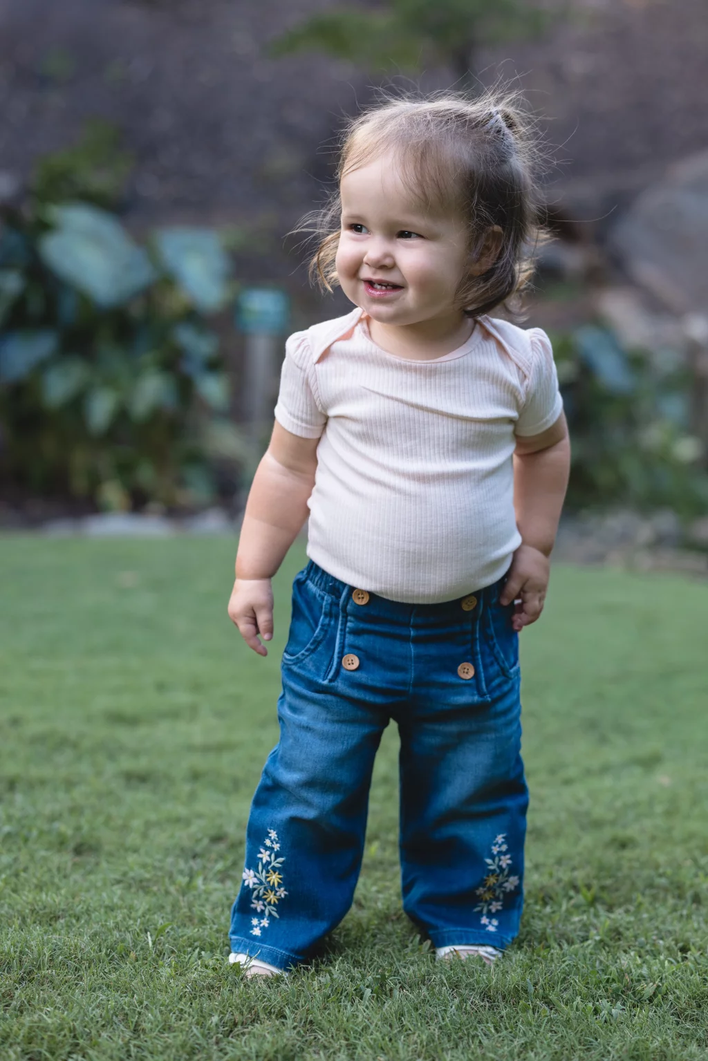 Family session at the Rock Quarry Garden.: Smiling toddler with light hair stands on a grassy lawn, wearing a pale pink shirt and embroidered blue jeans.