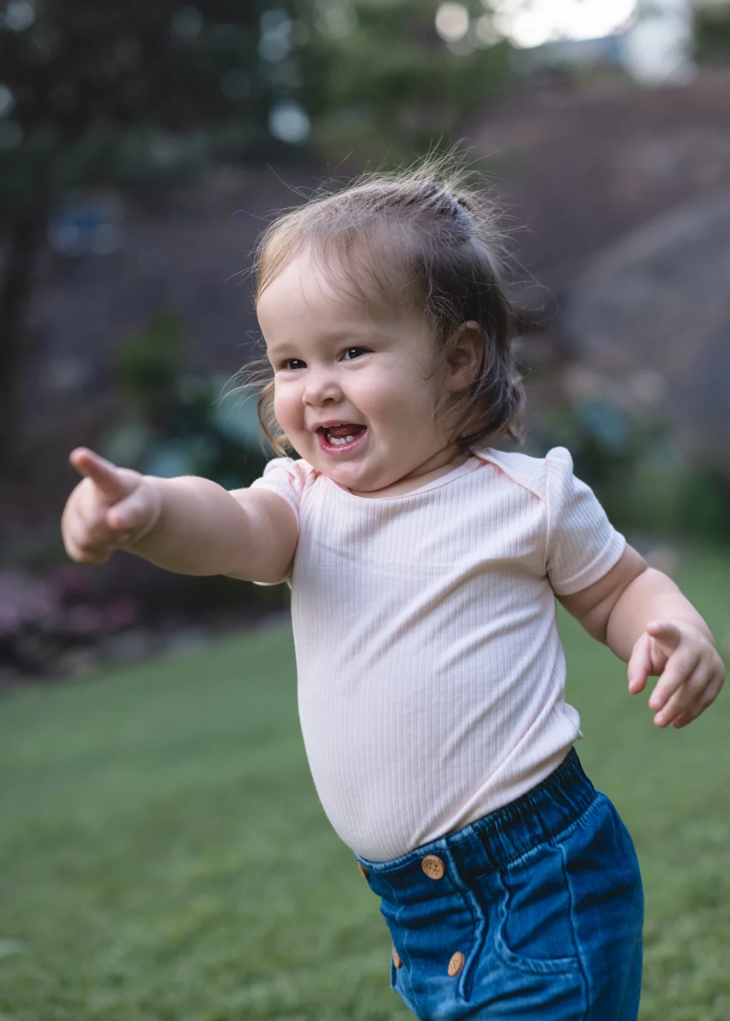 Family session at the Rock Quarry Garden.: Smiling toddler in a pale pink shirt and blue pants points excitedly while standing outdoors in a garden.