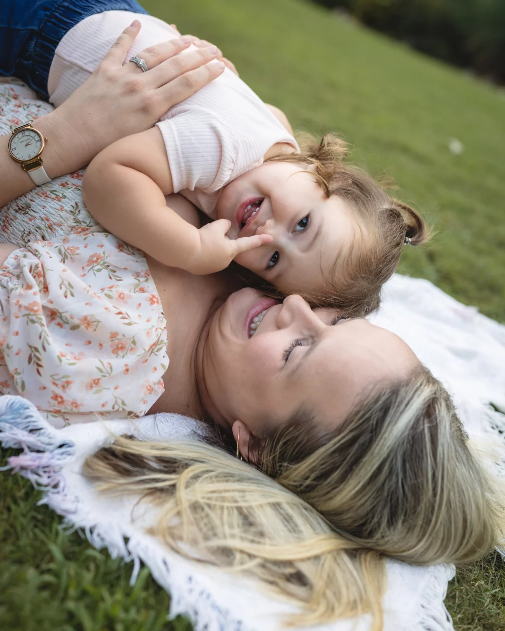 Family session at the Rock Quarry Garden.: A smiling child playfully hugs a woman lying on a blanket in the grass, with the woman&rsquo;s hand resting on the child&rsquo;s back.