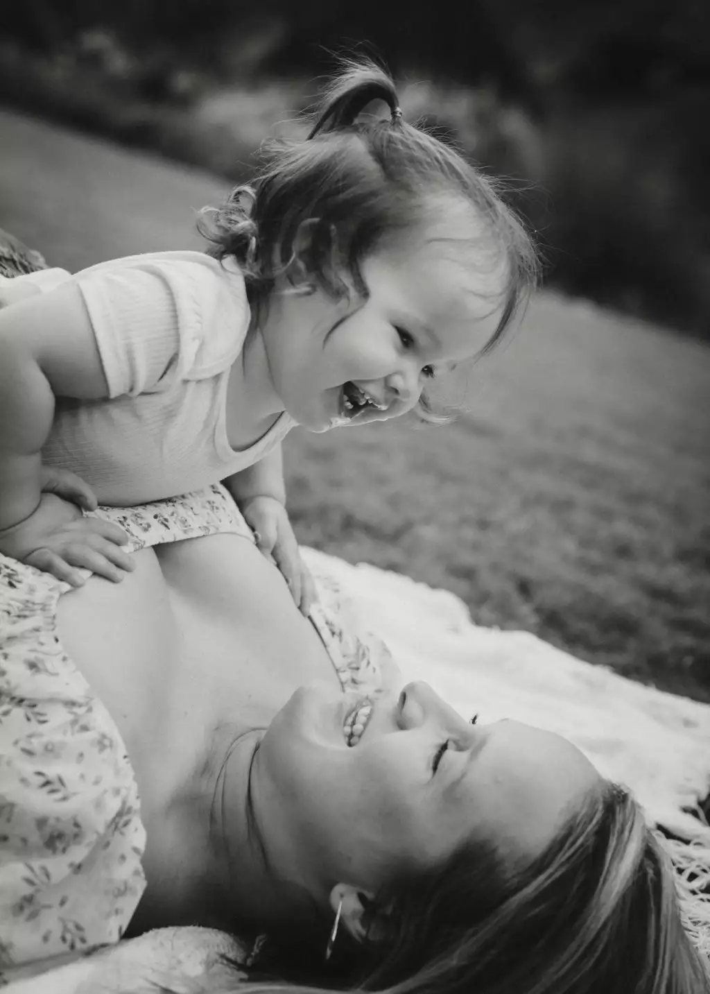 : Black-and-white photo of a smiling toddler leaning over and laughing with a woman lying on a blanket outdoors.