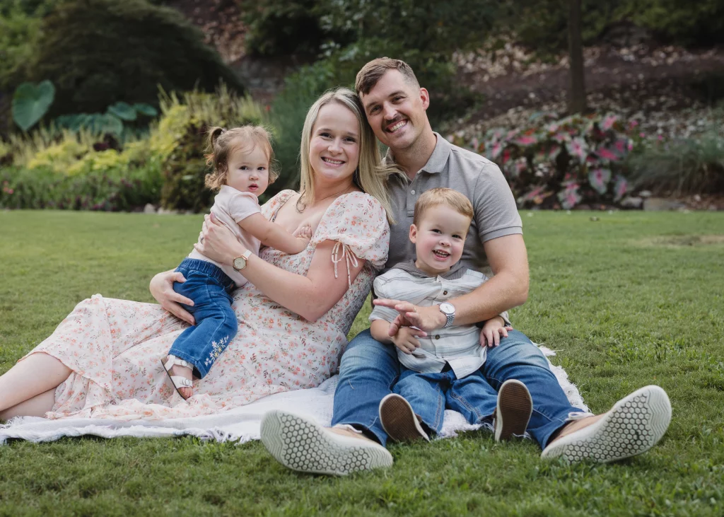 Family session at the Rock Quarry Garden.: A smiling family of four sits together on a blanket in a garden, with the mother holding a young daughter and the father holding their young son.