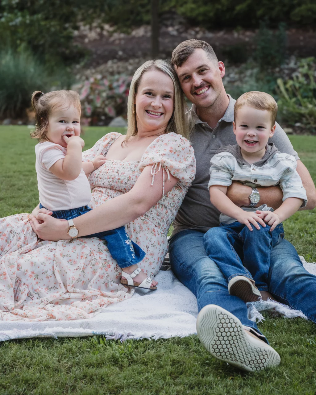 Family session at the Rock Quarry Garden.: Smiling family of four seated together on a picnic blanket in a grassy garden: parents with a young daughter and son.
