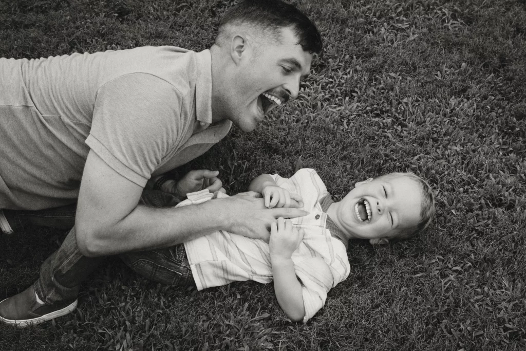 Family session at the Rock Quarry Garden.: Black-and-white photo of a smiling father playfully tickling a laughing young child lying on the grass.