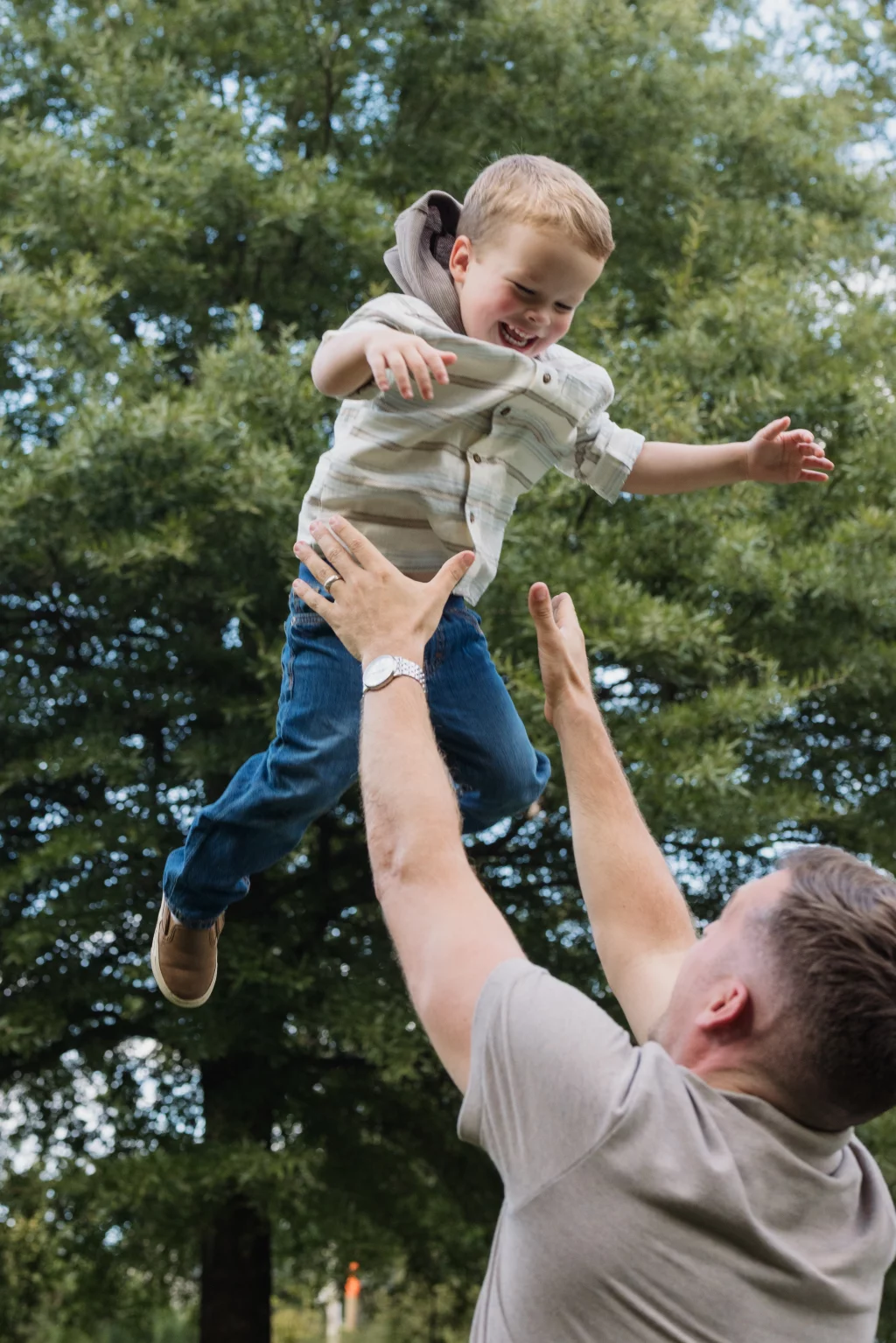 Family session at the Rock Quarry Garden.: A smiling young boy is lifted into the air by a man outdoors beneath leafy trees.