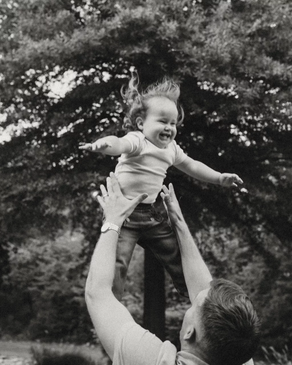 Family session at the Rock Quarry Garden.: Black-and-white outdoor photo of a smiling child being lifted into the air by an adult, with leafy trees in the background.