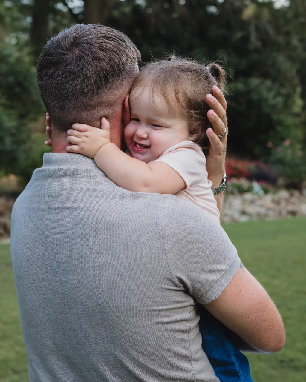 Family session at the Rock Quarry Garden.: A smiling toddler hugs a man from behind as he carries her outdoors in a garden.