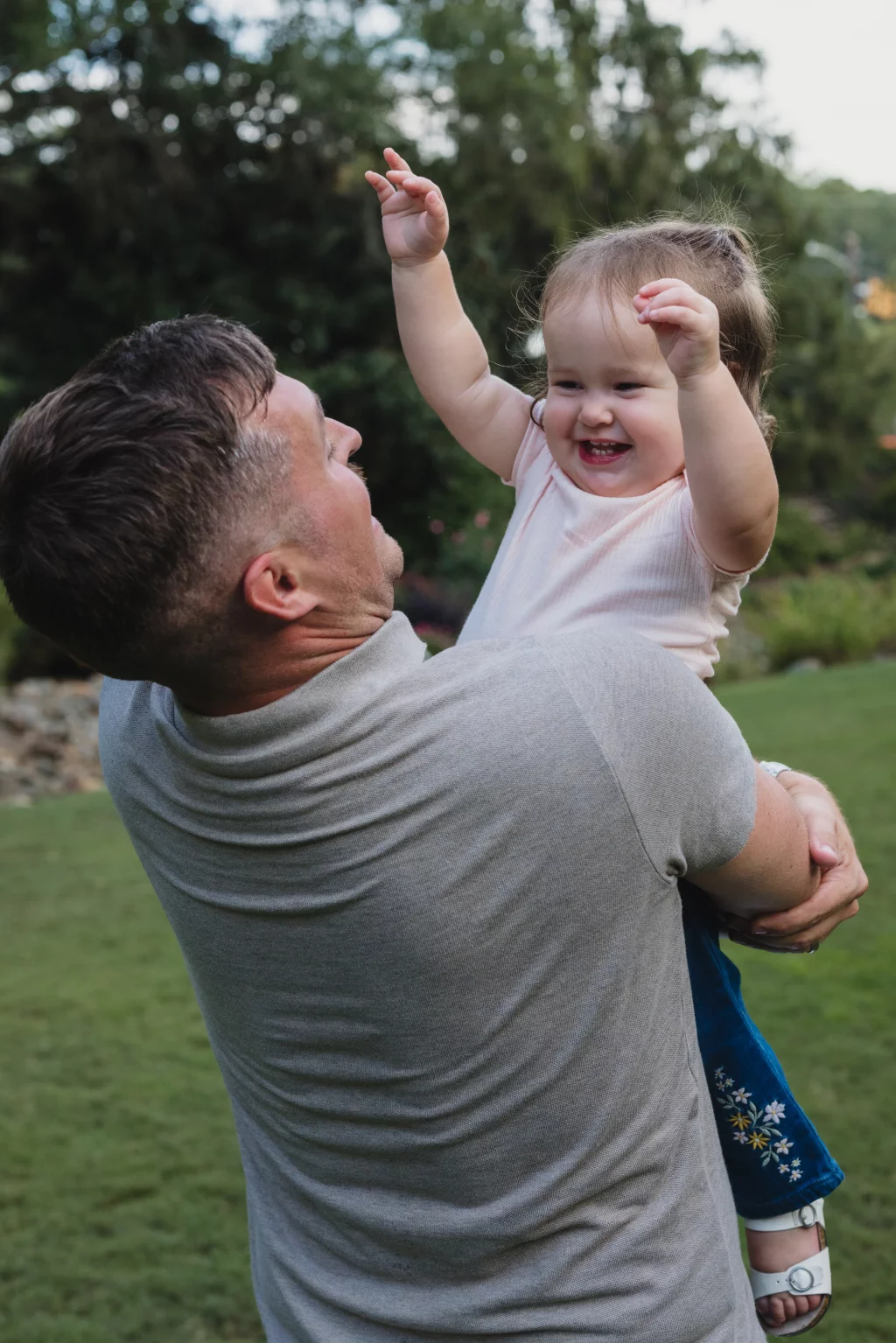 Family session at the Rock Quarry Garden.: A smiling toddler in a pink shirt and floral skirt raises both arms while being held outdoors by a man in a gray shirt.