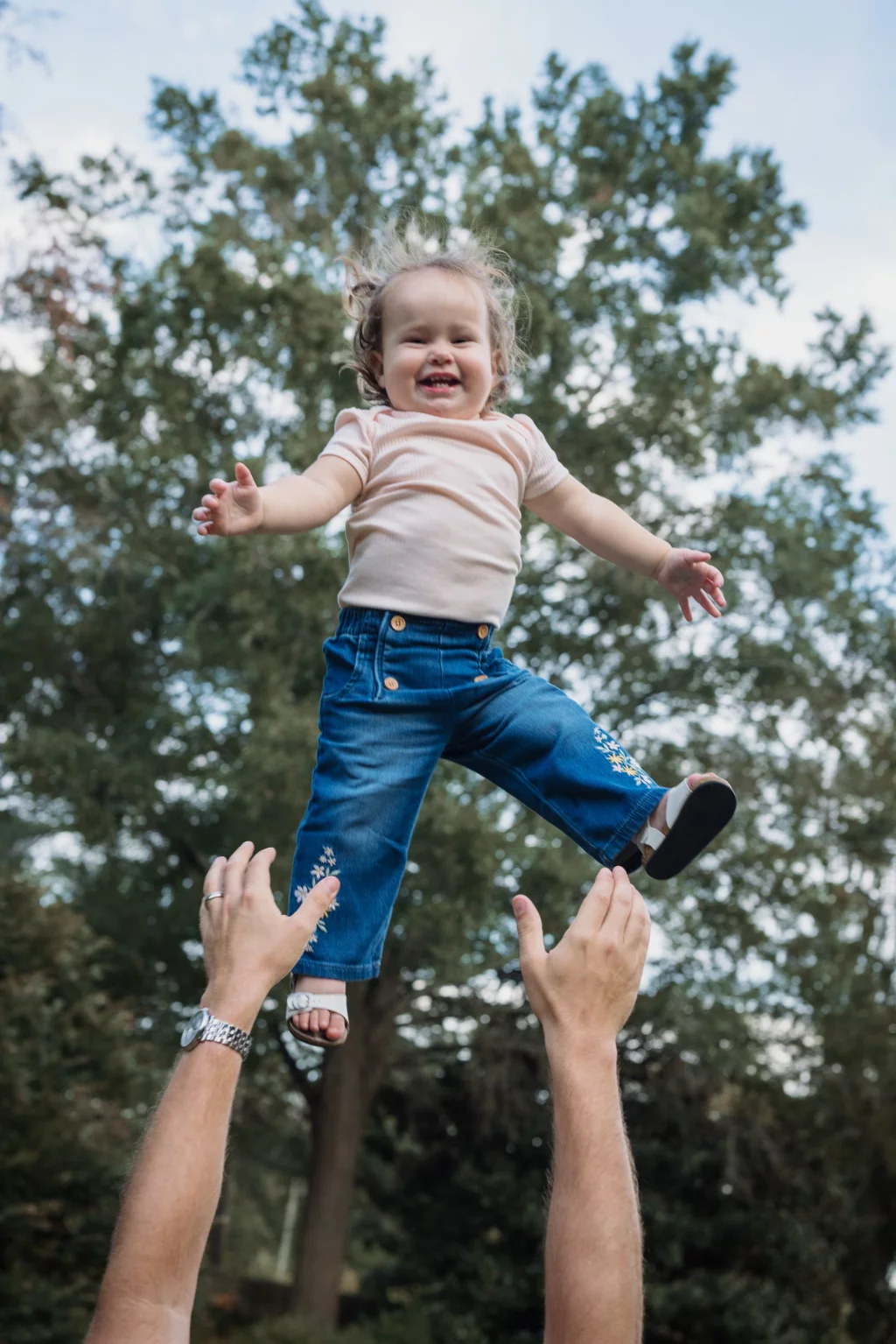 Family session at the Rock Quarry Garden.: Smiling toddler in a pink shirt and blue jeans is lifted into the air by an adult&rsquo;s outstretched hands in a tree-filled outdoor setting.