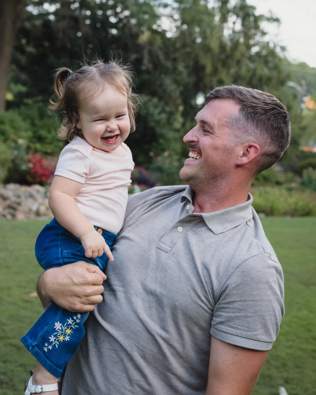 Family session at the Rock Quarry Garden.: A smiling man holds a laughing toddler in a garden. The child wears a pale shirt and embroidered blue skirt, while the man wears a gray polo shirt.