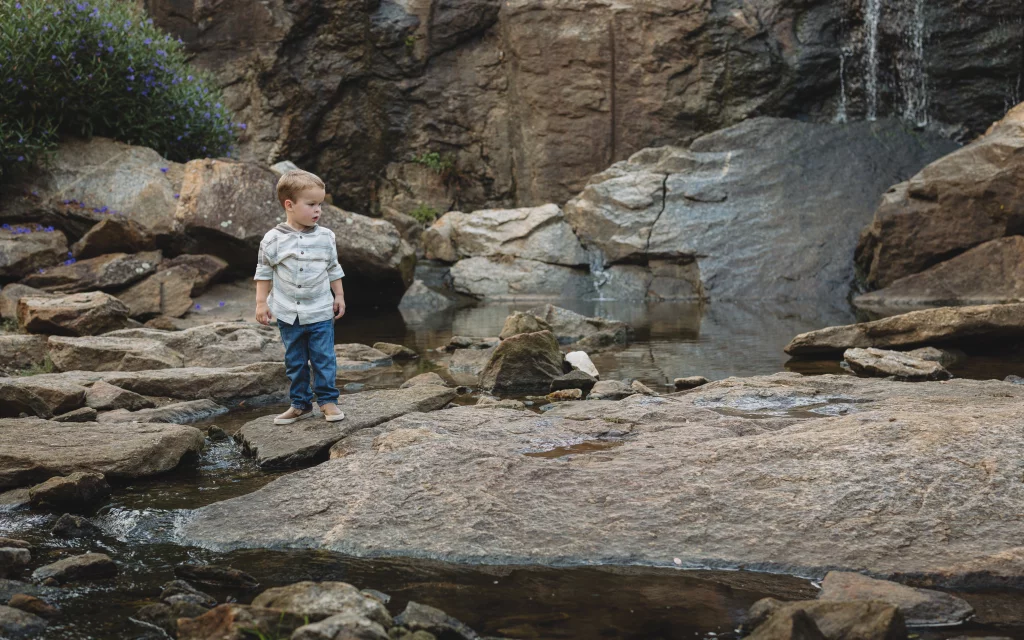 Family session at the Rock Quarry Garden.: Young boy standing on rocks beside a shallow stream and small waterfall, surrounded by a rocky gorge and purple wildflowers.