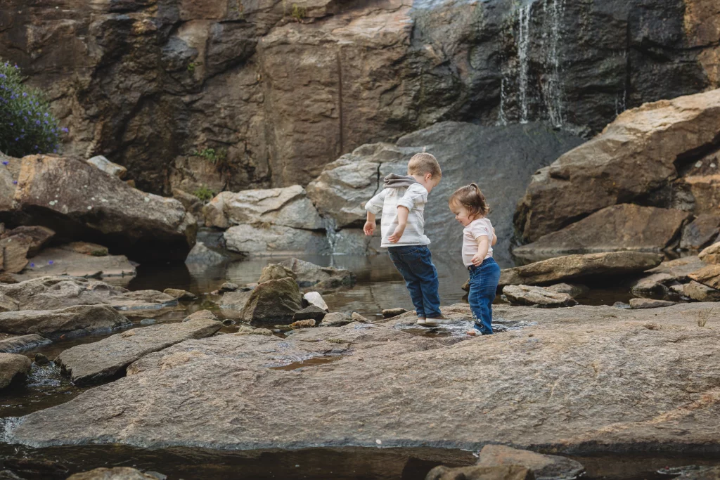Family session at the Rock Quarry Garden.: Two young children stand on rocks beside a shallow creek beneath a small waterfall.