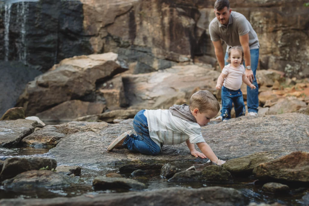 Family session at the Rock Quarry Garden.: A family explores a rocky creek beneath a waterfall, with a young boy crawling through shallow water and a man helping a toddler stand on the rocks.