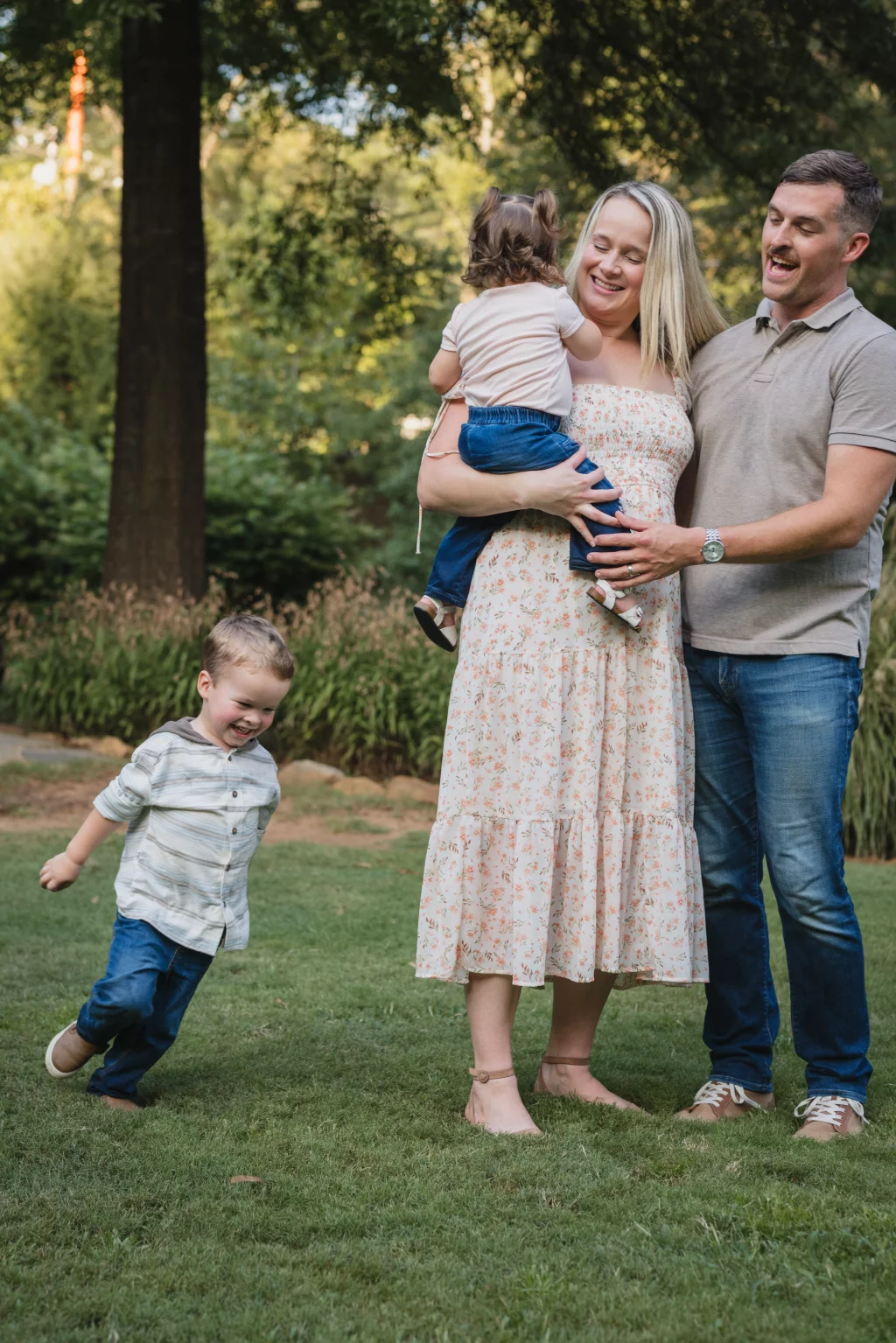 Family session at the Rock Quarry Garden.: A smiling family of four poses outdoors in a wooded park: the mother holds a toddler, the father stands beside them, and a young boy runs across the grass.