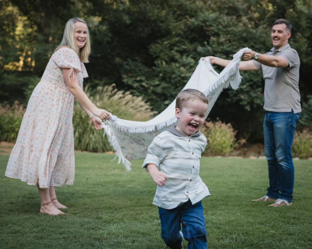 Family session at the Rock Quarry Garden.: A laughing child runs across a lawn while two smiling adults hold and swing a fringed blanket behind him in a wooded garden.