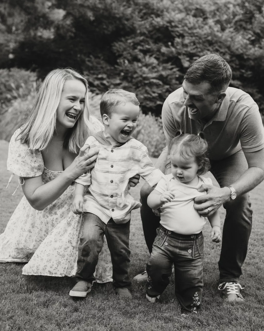 Family session at the Rock Quarry Garden.: Black-and-white family portrait outdoors, with two smiling parents playfully supporting their laughing young son and daughter on a grassy lawn.