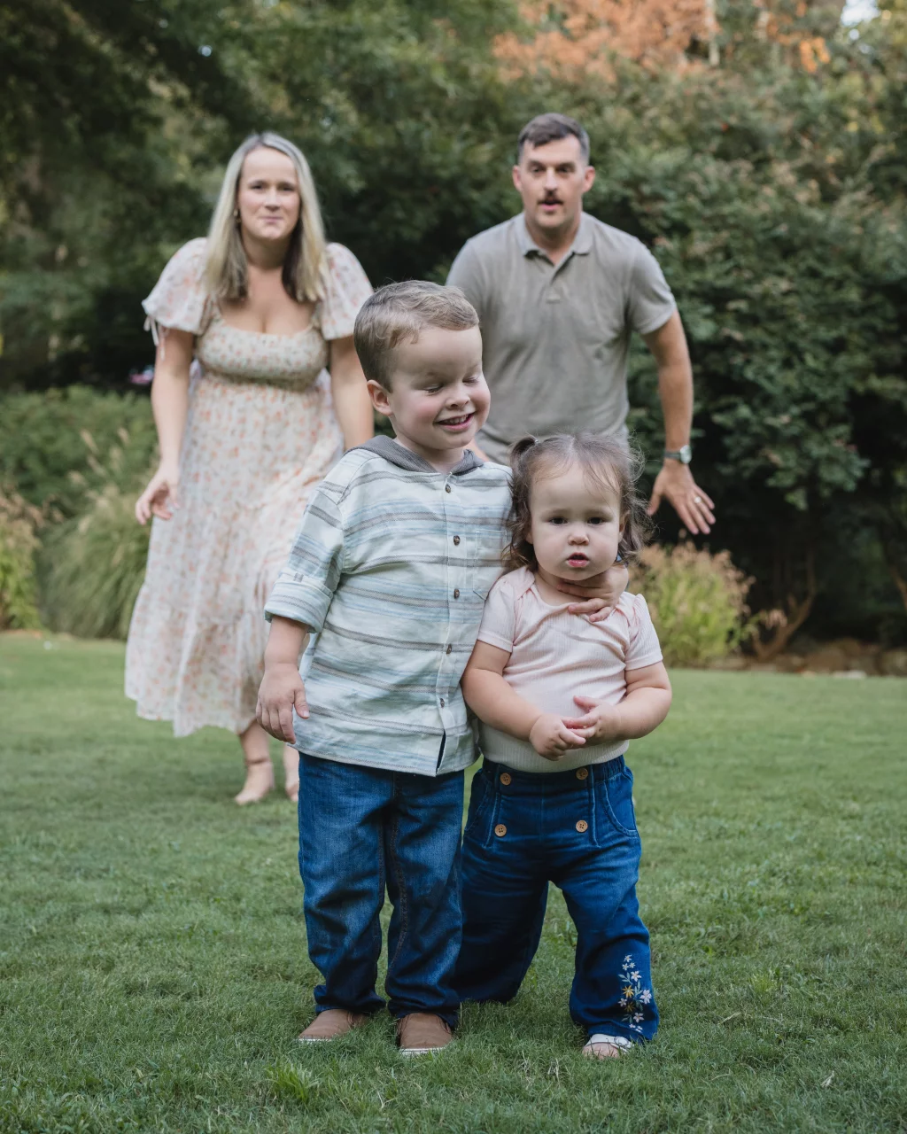 Family session at the Rock Quarry Garden.: A family of four poses on a grassy lawn, with two young children in front and their parents standing behind them amid trees.