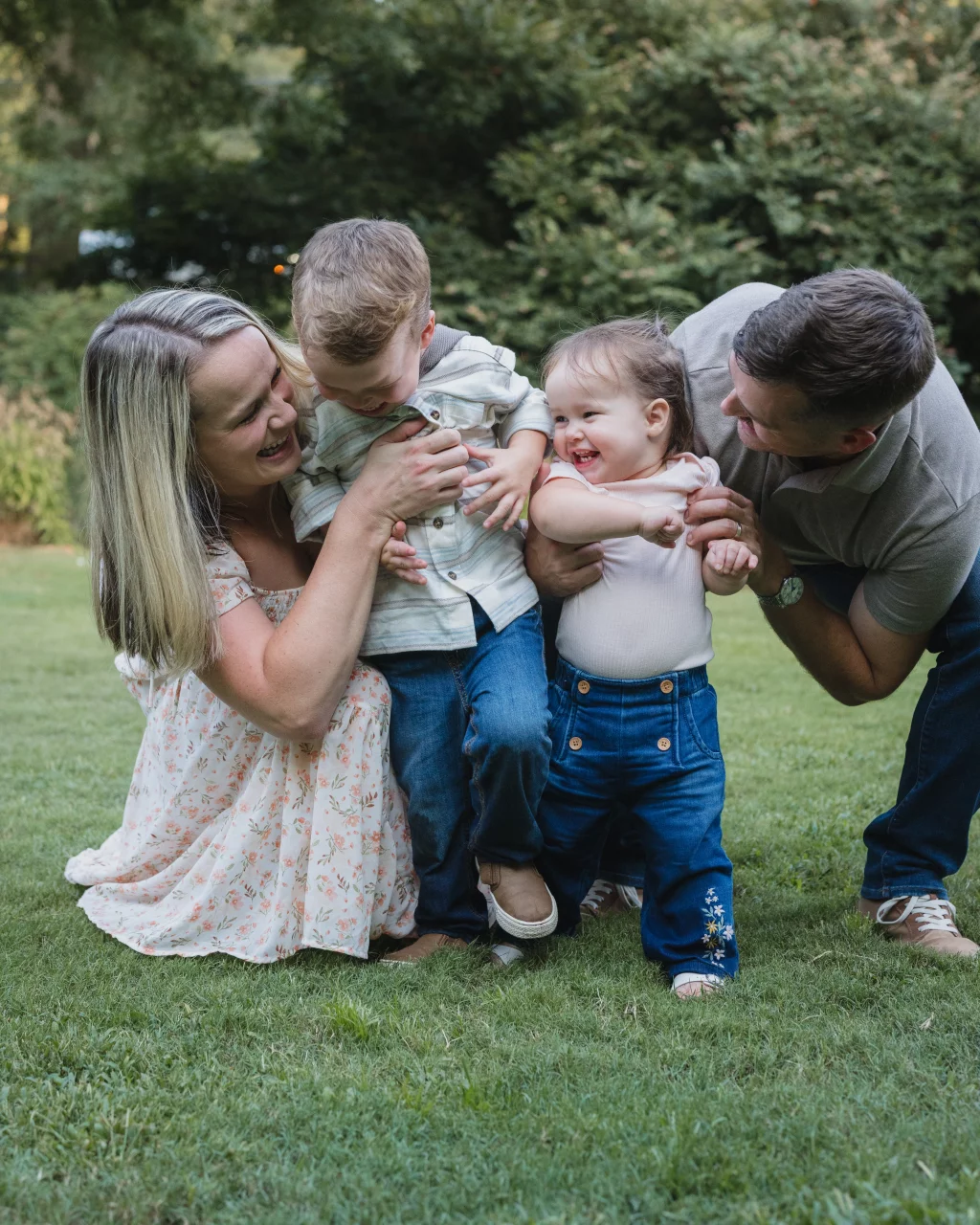 Family session at the Rock Quarry Garden.: Smiling parents play with their two young children on a lawn in a leafy park.