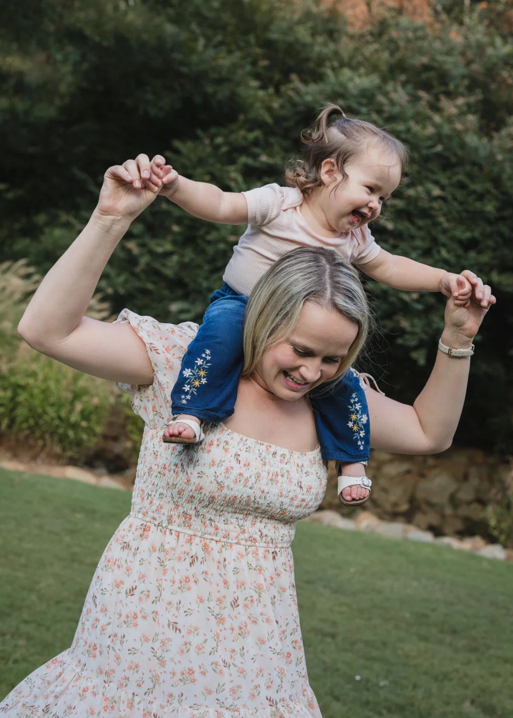 Family session at the Rock Quarry Garden.: A smiling woman in a floral dress carries a laughing toddler on her shoulders outdoors, holding the child&rsquo;s hands as they play.