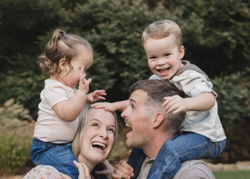 Family session at the Rock Quarry Garden.: Smiling family outdoors, with a father carrying a young boy on his shoulders and a mother carrying a young girl, surrounded by trees.