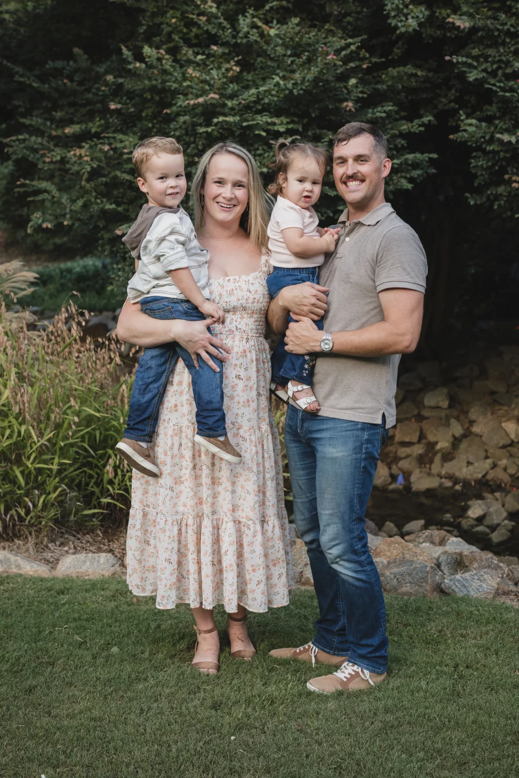 Family session at the Rock Quarry Garden.: A smiling family of four poses outdoors on a lawn beside a rocky stream, with the parents holding two young children in front of dense greenery.