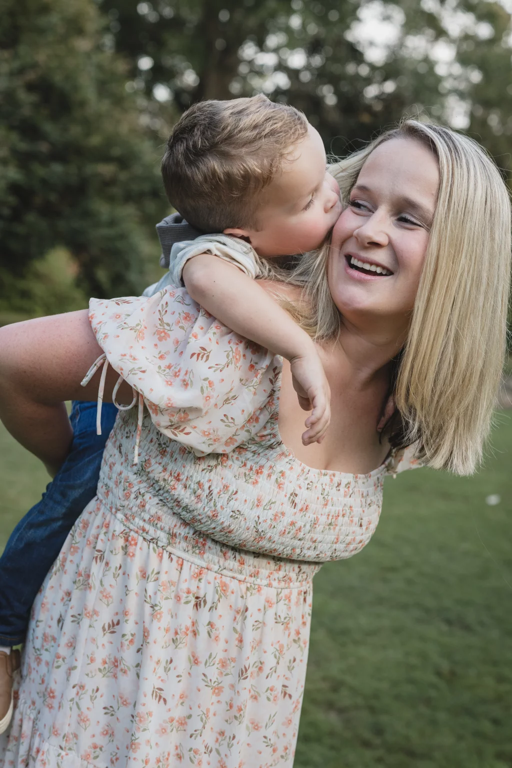 Family session at the Rock Quarry Garden.: A smiling woman in a floral dress holds a young child, who kisses her cheek, outdoors in a wooded park.