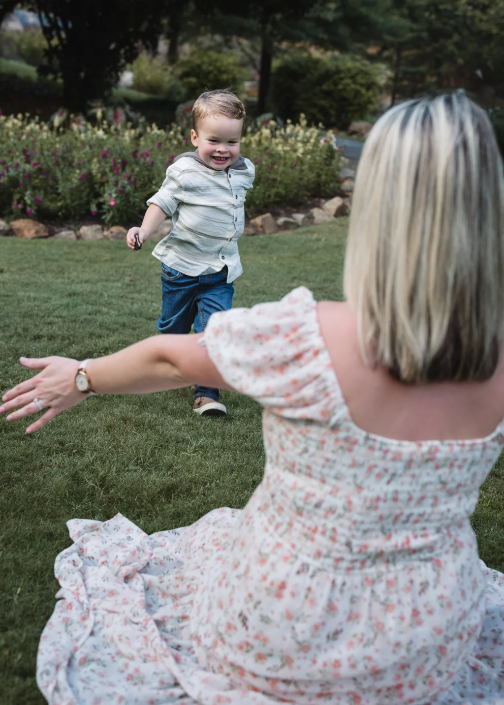 Family session at the Rock Quarry Garden.: A smiling young boy runs across a lawn toward a woman in a floral dress with her arms outstretched, surrounded by a flower garden.