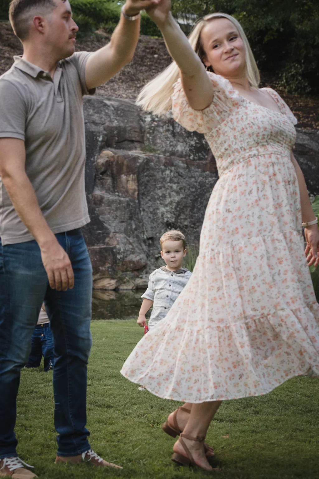 Family session at the Rock Quarry Garden.: A smiling pregnant woman in a floral dress twirls outdoors while holding hands with a man; a young boy stands behind them near a stone wall.
