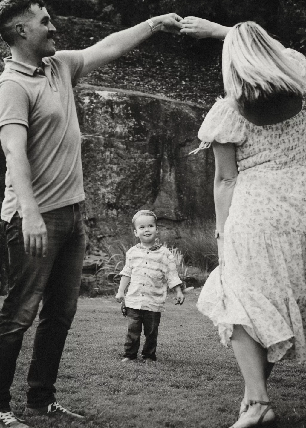 Family session at the Rock Quarry Garden.: Black-and-white outdoor photo of a smiling man and woman holding hands overhead as if dancing, with a young boy standing between them on the grass.
