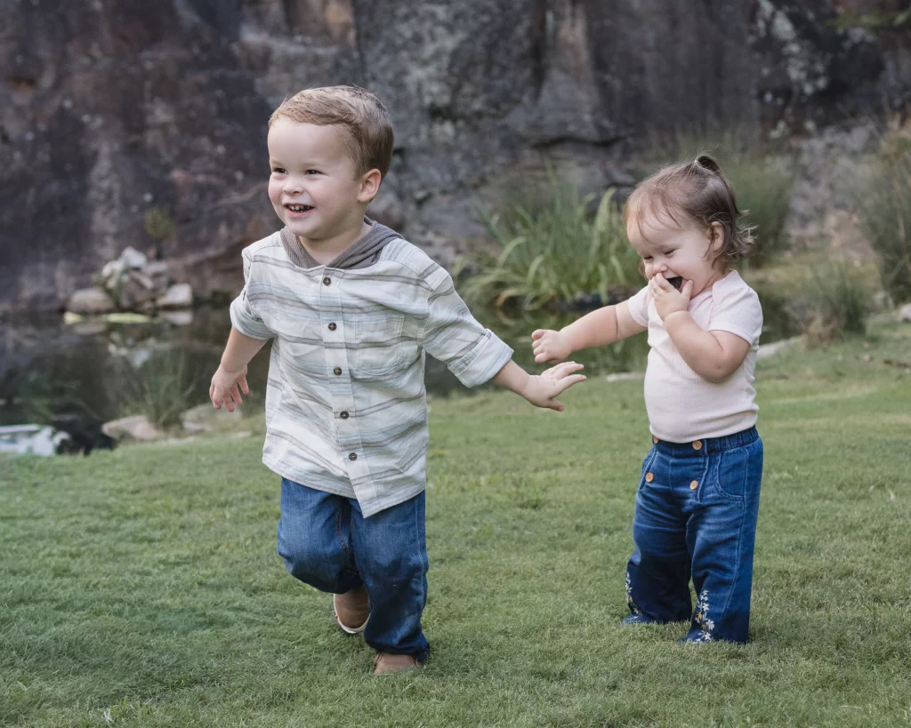 Family session at the Rock Quarry Garden.: Two young children play on a grassy lawn beside a small pond and rocky garden; the boy runs smiling while the girl stands nearby with a hand to her mouth.