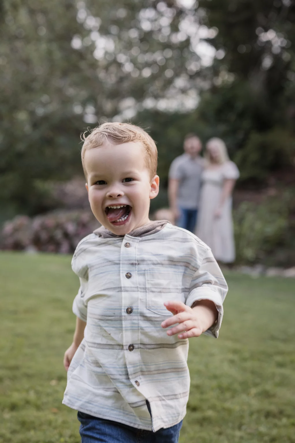Family session at the Rock Quarry Garden.: A smiling young boy in a striped shirt runs toward the camera on a lawn, with two adults blurred in the wooded background.