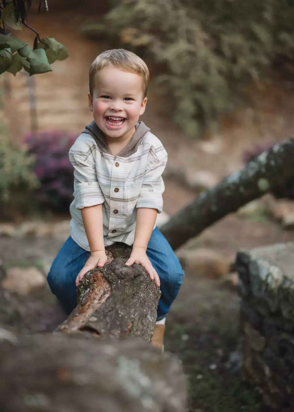 : Smiling young boy sitting on a fallen log outdoors, wearing a striped shirt and blue jeans, surrounded by blurred greenery and rocks.