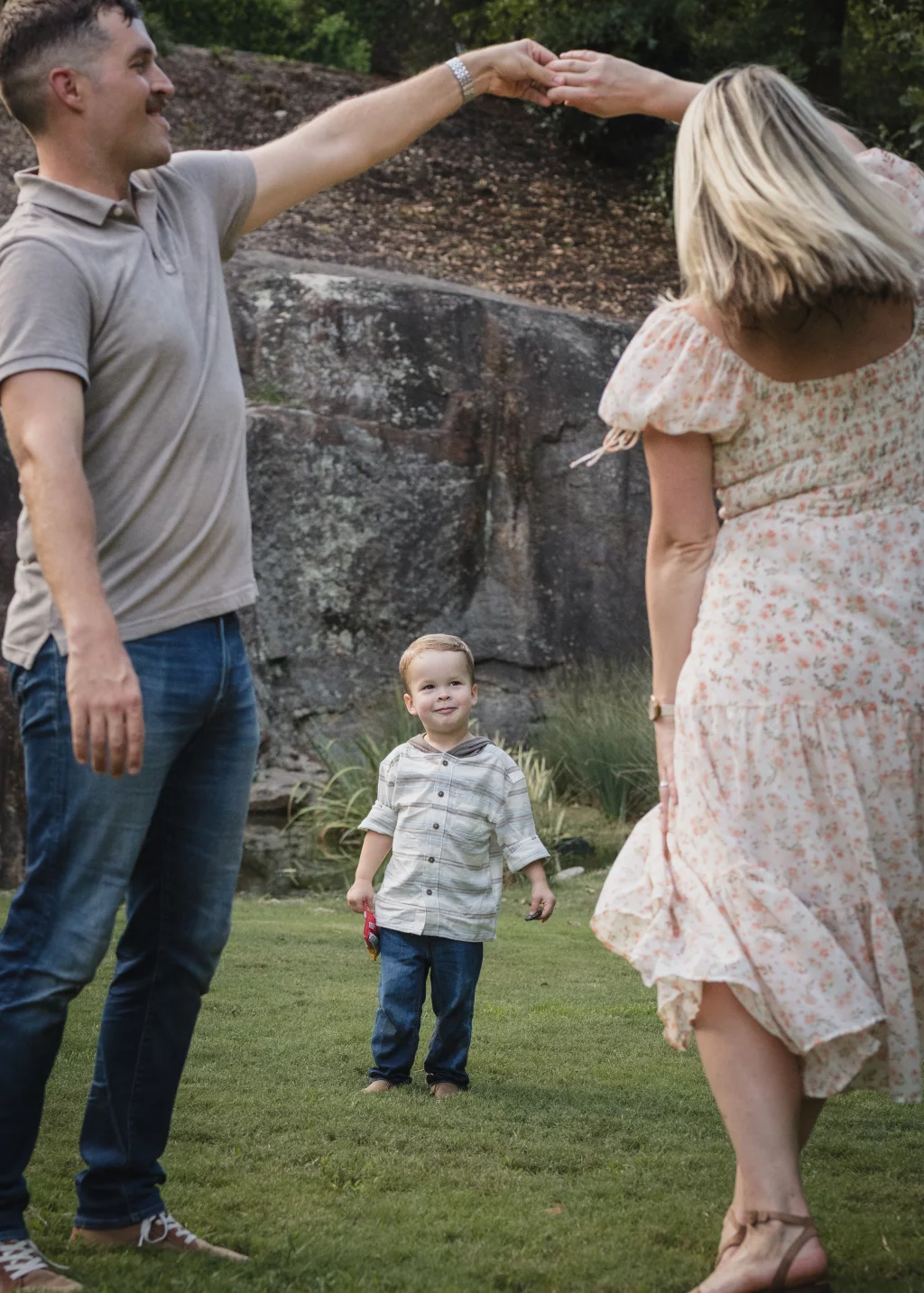Family session at the Rock Quarry Garden.: A man and woman playfully touch hands overhead while their young son stands between them on a grassy lawn near a stone wall.