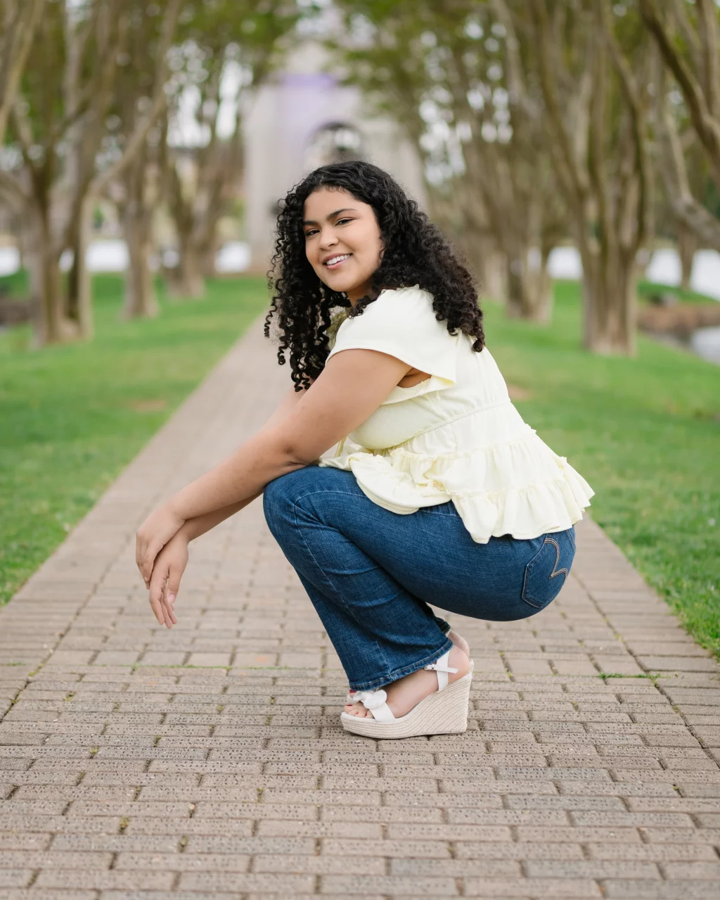 Nolan's senior photos taken at Furman University: Woman with curly hair squatting on a brick walkway, wearing a light yellow top, blue jeans, and wedge sandals, with trees lining the path in the background.