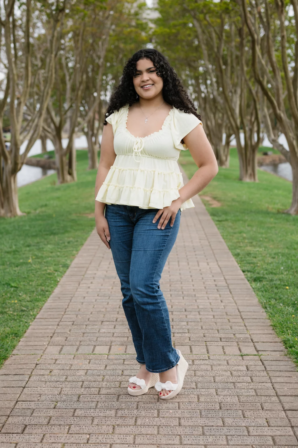 Nolan's senior photos taken at Furman University: Woman with curly hair wearing a pale yellow blouse and blue jeans stands on a brick path lined with trees and grass, smiling at the camera.