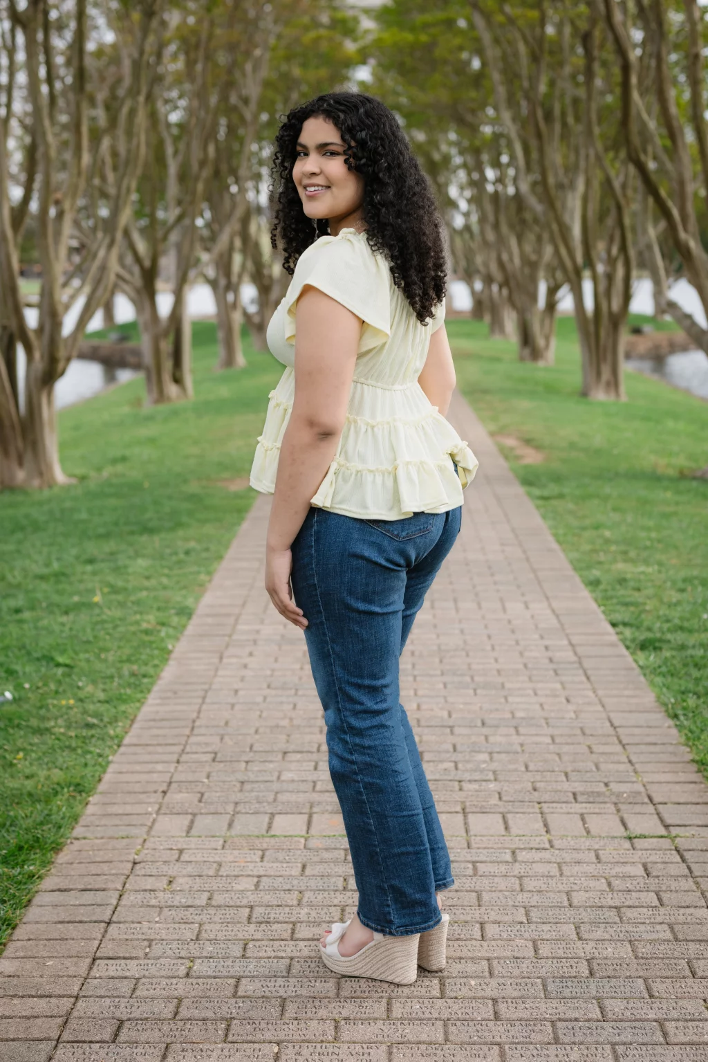 Nolan's senior photos taken at Furman University: A woman with curly hair wearing a pale yellow ruffled top, blue jeans, and wedge sandals stands on a brick path in a park, looking back and smiling. Trees line both sides of the path.